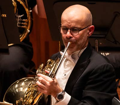 Man in a tuxedo playing a French horn in an orchestra setting.