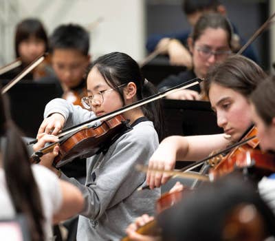 Students playing violins in an orchestra, focused and engaged in their performance.