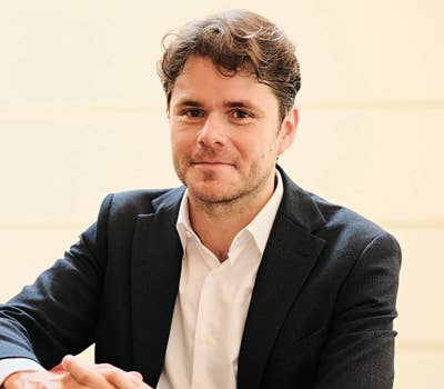 Man in a dark suit and white shirt sitting with hands clasped, smiling slightly against a light background.