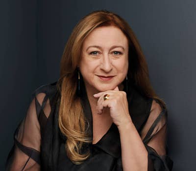 Woman with long hair and sheer black top, resting her chin on her hand, against a dark gray background.