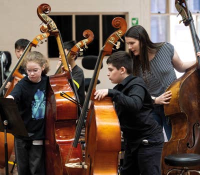 Children playing double basses with guidance from an instructor in a music classroom setting.